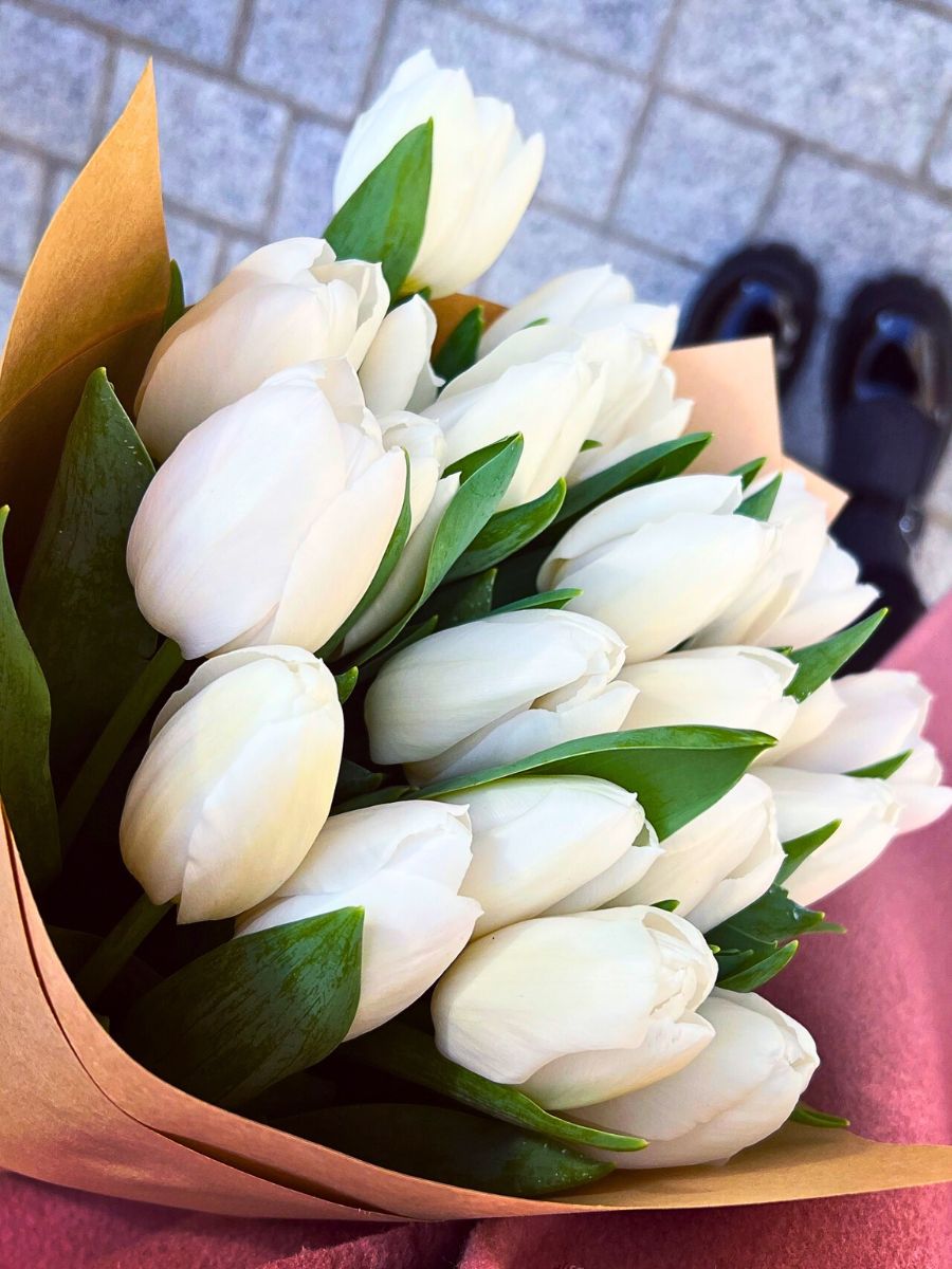 A bouquet of fresh white tulips wrapped in brown paper, held against a blurred stone pavement with black shoes visible in the background.