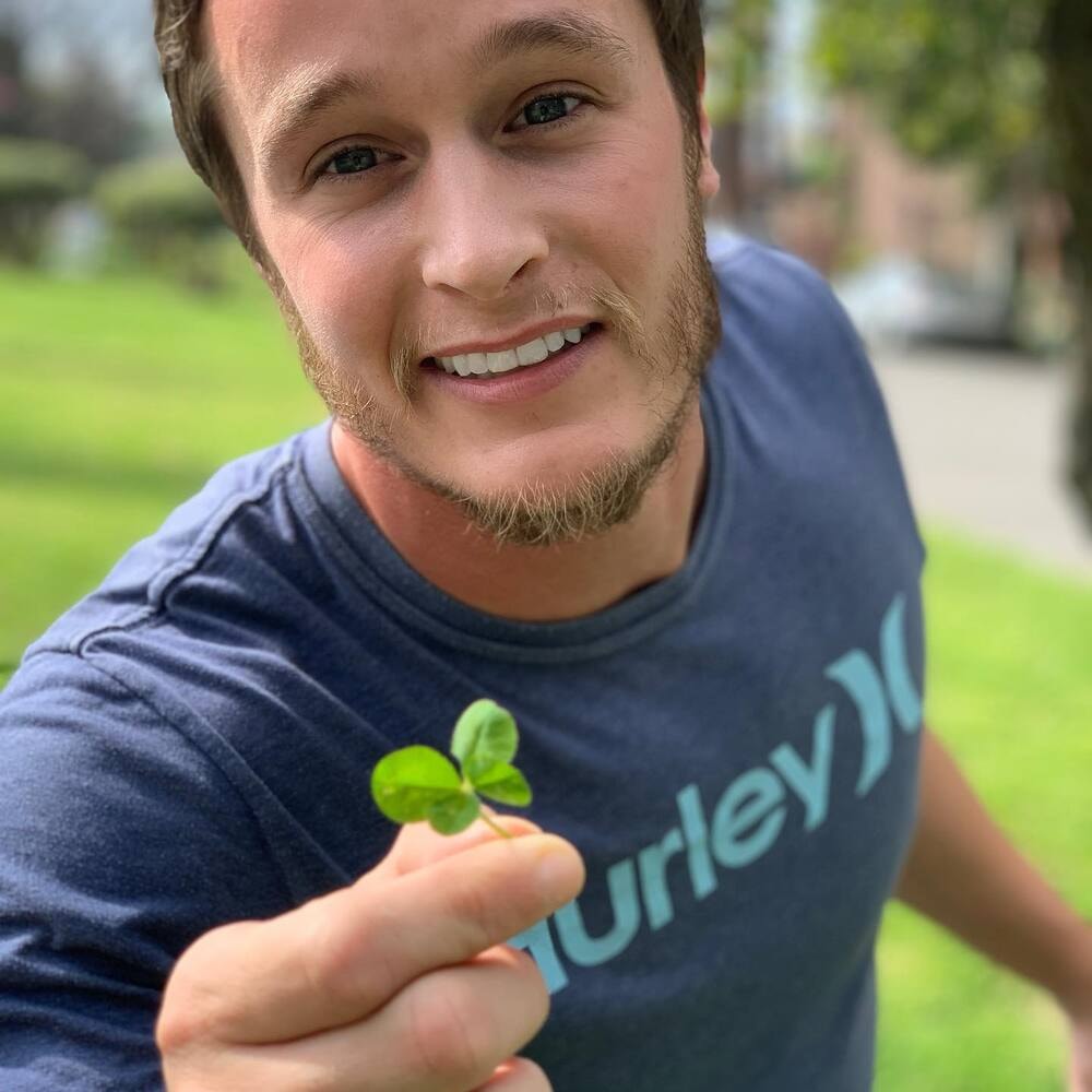 A smiling man showing a small clover while standing in a green area